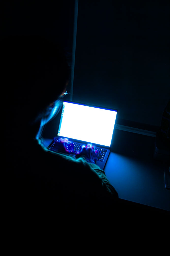Photo of boy typing on a laptop in a dark room, lit only by his laptop screen