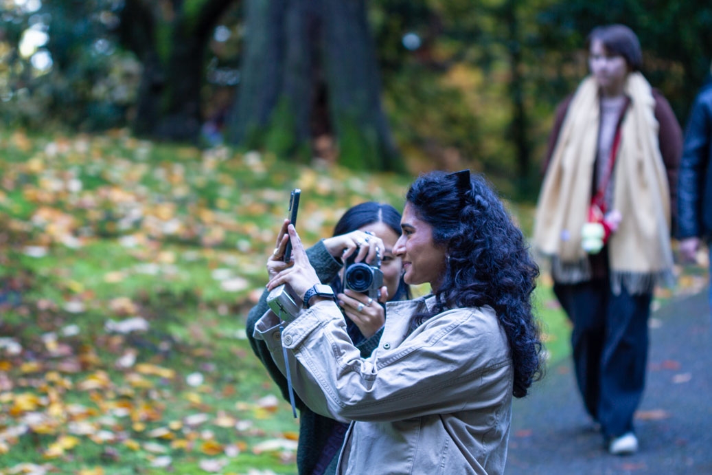 Picture of two friends, one facing left taking a picture with a smartphone, and the other facing the viewer with a camera